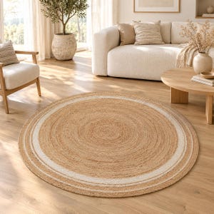 May include: A round area rug crafted from natural tan jute fibers, featuring a concentric circular pattern with a distinct white border ring. The rug has a textured, woven surface and is laid flat on a light-colored wooden floor.