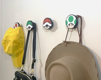 May include: Three mushroom-shaped hooks with different colored tops, green, red, and yellow, are mounted on a white wall. A yellow baseball cap, a black purse, and a brown cowboy hat are hanging from the hooks.