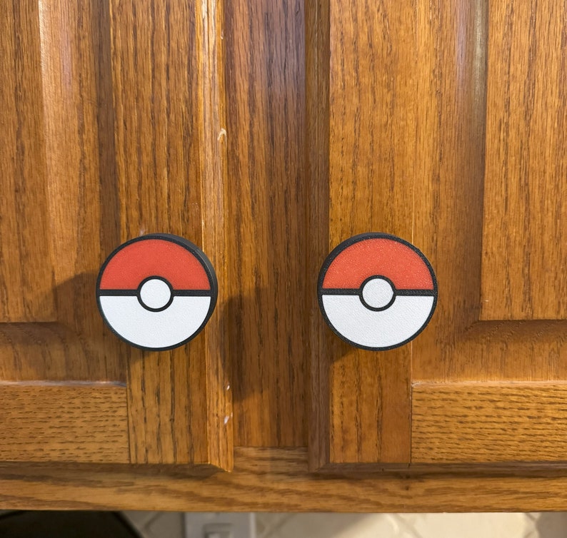 Puede incluir: Dos tiradores de armario con un dise&ntilde;o rojo y blanco que se asemeja a una Pok&eacute; Ball. Los tiradores tienen un borde negro y un c&iacute;rculo blanco en el centro. Montados en un armario de madera.