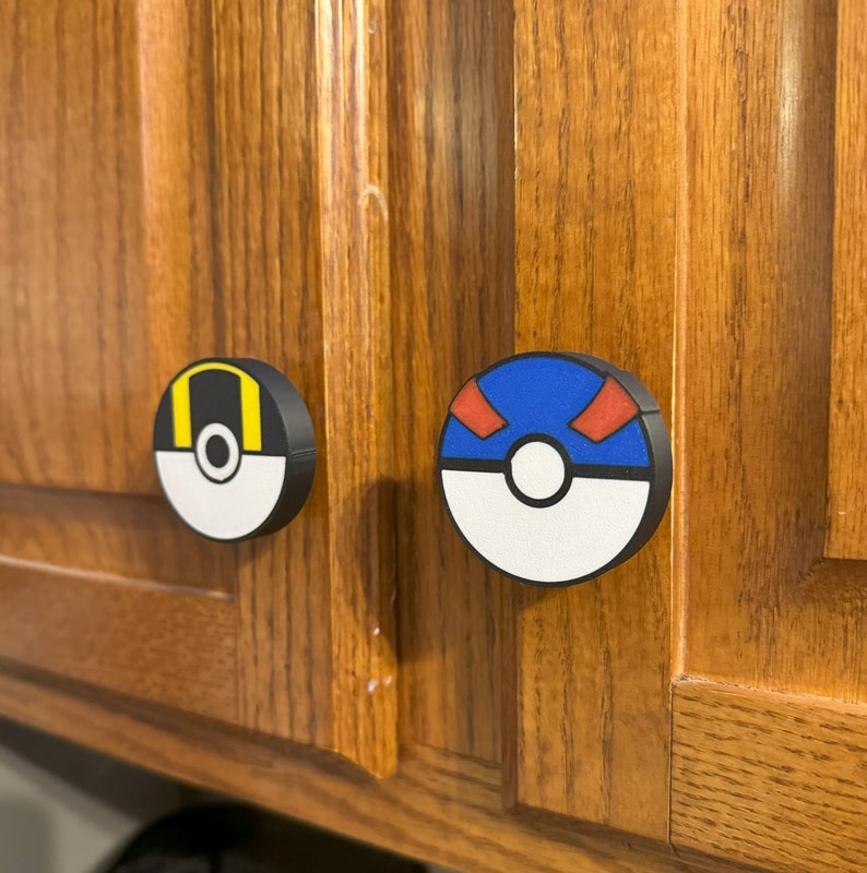 Puede incluir: Dos pomos de armario redondos con un dise&ntilde;o negro, blanco, amarillo, azul y rojo. Los pomos presentan un dise&ntilde;o de dibujos animados. Los pomos est&aacute;n montados en un armario de madera.