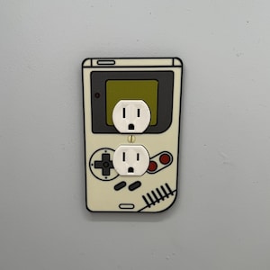Peut inclure: Une plaque murale de style Game Boy blanche et noire avec deux prises électriques. Les prises sont blanches et ont un centre noir.
