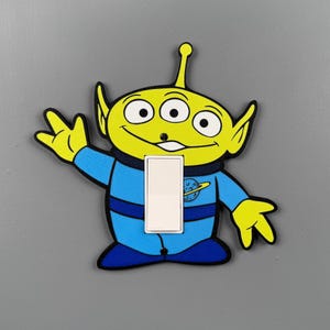 May include: A light switch cover shaped like a three-eyed alien character with lime green skin and a blue space suit. The character has a wide smile, pointed ears, and an antenna. A white rectangular rocker switch is centered on the alien's torso.
