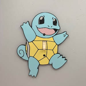 May include: A light switch cover featuring a cartoon Squirtle from the Pokemon franchise. The cover is made of wood and painted in blue, yellow, and black. The switch is white and is located on the Squirtle's shell.