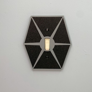 May include: A black and gray light switch cover with a hexagonal design featuring a Star Wars TIE Fighter silhouette.