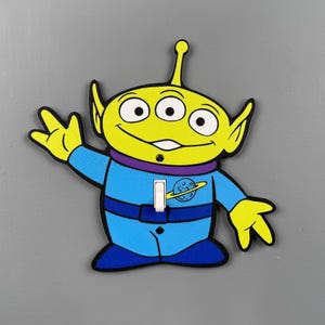 May include: A light switch cover shaped like a three-eyed alien character with a bright yellow head, pointed ears, and a blue space suit. The suit features a purple collar, a dark blue belt, and a small planet graphic on the chest area.