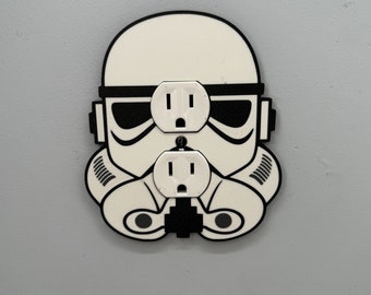 May include: A white and black wall outlet cover shaped like a Star Wars Stormtrooper helmet. The outlet cover has two standard electrical outlets.