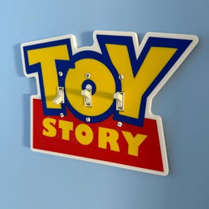 May include: A light switch cover featuring the "Toy Story" logo in yellow, blue, and red. The cover has three light switches and is mounted on a light blue wall. The word "TOY" is above "STORY".