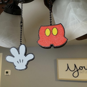 May include: Two decorative fan pull chains featuring a white hand and red shorts with yellow ovals, reminiscent of a famous cartoon character. The pulls hang from a ceiling fan with white glass shades. A sign with the word "You" is visible.
