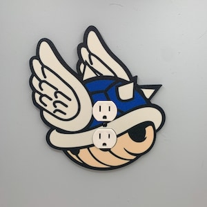 May include: A blue and white wall outlet cover shaped like a winged turtle shell from the Super Mario Bros. video game series.