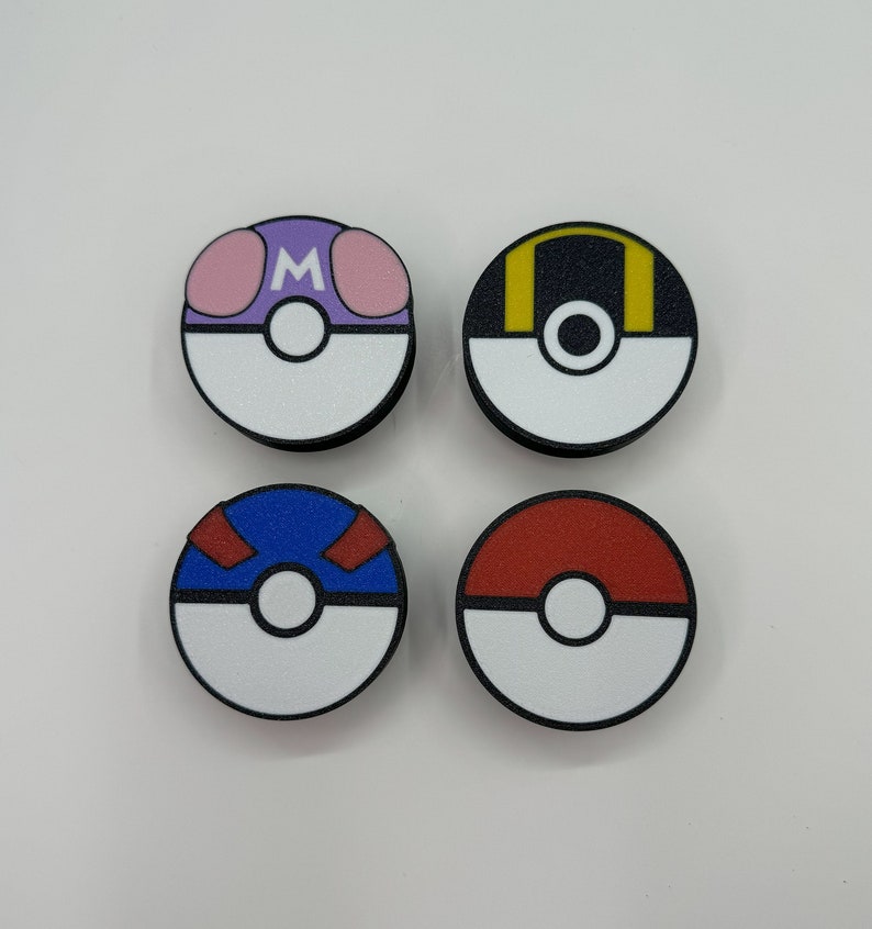 Puede incluir: Cuatro dise&ntilde;os circulares y coloridos que se asemejan a Pok&eacute; Balls. Cada bola presenta una mitad inferior blanca y un borde negro. Las mitades superiores son moradas con acentos rosas y una "M" blanca, negras con acentos amarillos, azules con acentos rojos y rojas s&oacute;lidas.
