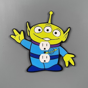 May include: A decorative wall outlet cover shaped like a three-eyed green alien character wearing a blue space suit with a purple collar. The design features a small planet graphic on the chest and two white electrical socket openings centered.