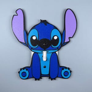 May include: A blue and purple light switch cover featuring a cartoon Stitch character from the Disney movie Lilo & Stitch. The character is sitting with its arms crossed and has a big smile.