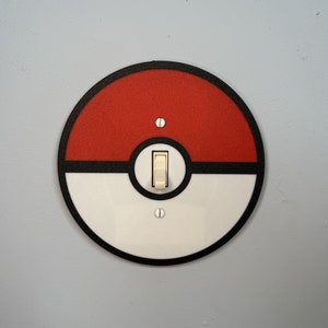 May include: A round light switch cover with a black border and a red and white Pokeball design.