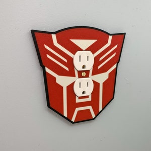 Peut inclure: Une plaque de prise électrique rouge et blanche sur le thème des Transformers avec deux prises standard.