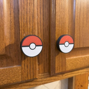 Puede incluir: Dos tiradores de armario con un dise&ntilde;o de Pokeball rojo y blanco. Los tiradores redondos tienen un borde negro y est&aacute;n montados en una puerta de armario de madera. Estos tiradores ofrecen un toque divertido y funcional a cualquier habitaci&oacute;n.