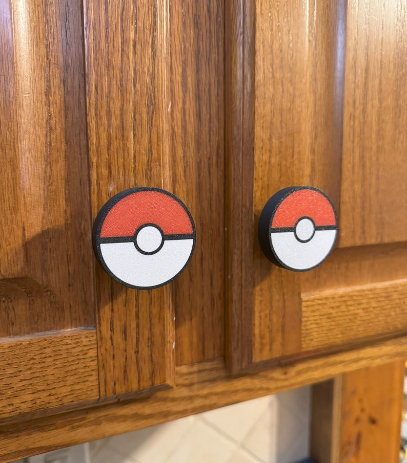Puede incluir: Dos tiradores de armario con un dise&ntilde;o de Pokeball rojo y blanco. Los tiradores redondos tienen un borde negro y est&aacute;n montados en una puerta de armario de madera. Estos tiradores ofrecen un toque divertido y funcional a cualquier habitaci&oacute;n.