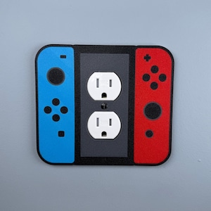 Peut inclure: Une plaque de prise de courant bleue et rouge avec un design Nintendo Switch. La plaque comporte deux prises électriques standard et une image stylisée des boutons et des manettes de la console Switch.