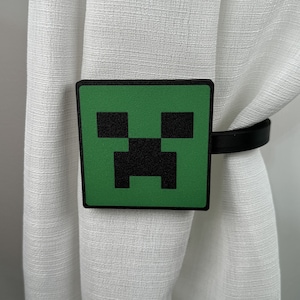 以下が含まれることがあります： 緑と黒のMinecraftクリーパーキャラクターのカーテン留め。カーテン留めはプラスチック製で、黒いストラップが付いています。