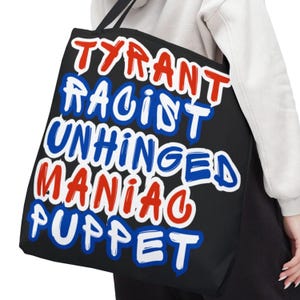 Puede incluir: Bolso tote negro con las palabras "TYRANT RACIST UNHINGED MANIAC PUPPET" en letras rojas, blancas y azules. El bolso tiene asas negras. Una persona sostiene el bolso, vistiendo una sudadera blanca.
