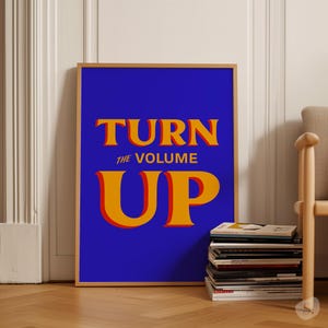 Puede incluir: Una impresión artística enmarcada con un fondo azul y las palabras "TURN THE VOLUME UP" en naranja con una sombra roja. La impresión está en un marco de madera y está apoyada contra una pared junto a una pila de libros.