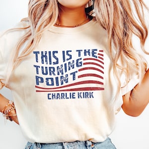 May include: Cream-colored t-shirt featuring the text "THIS IS THE TURNING POINT" in blue, with red and white stripes, and the name "CHARLIE KIRK" below. The shirt is made of a soft material.