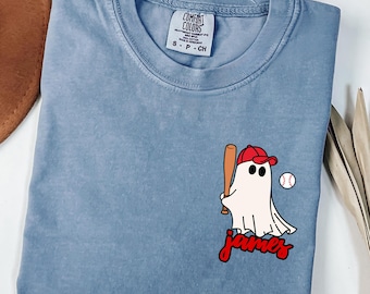 May include: A light blue t-shirt with a cartoon ghost wearing a red baseball cap and holding a bat. A baseball is next to the ghost, and the name "James" is written in red script below the ghost.