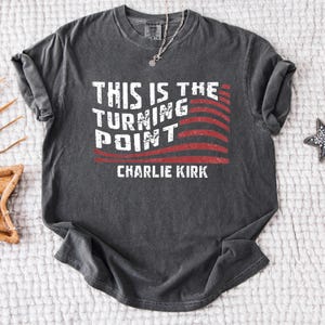 May include: Dark gray t-shirt with the text "THIS IS THE TURNING POINT" in white, with a red and white striped design. The name "CHARLIE KIRK" is printed below the text. A silver necklace is visible.