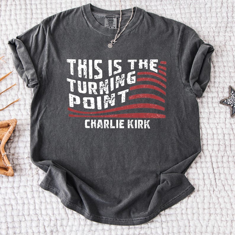 May include: Dark gray t-shirt with the text "THIS IS THE TURNING POINT" in white, with a red and white striped design. The name "CHARLIE KIRK" is printed below the text. A silver necklace is visible.