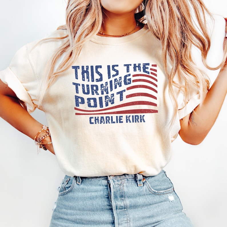 May include: Cream-colored t-shirt featuring the text "THIS IS THE TURNING POINT" in blue, with red and white stripes, and the name "CHARLIE KIRK" below. The shirt is made of a soft material.
