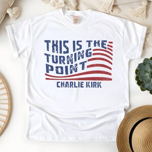 May include: White t-shirt with the text "THIS IS THE TURNING POINT" in blue, with a red and white striped design, and the name "CHARLIE KIRK" in blue. The shirt is a casual wear item.