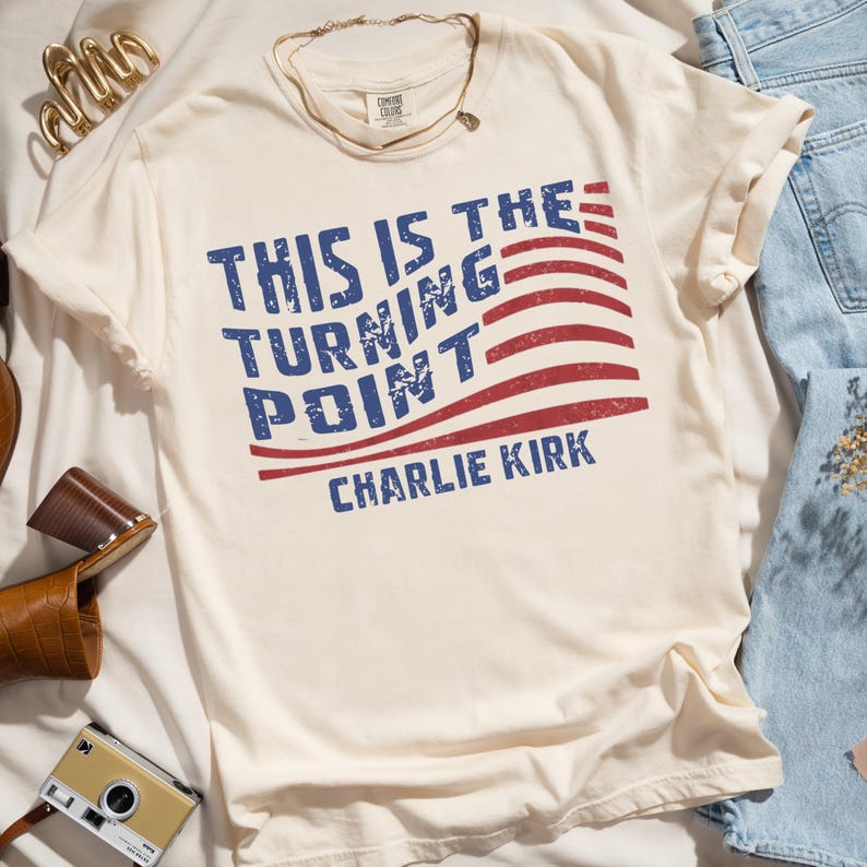 May include: Cream-colored t-shirt with the text "THIS IS THE TURNING POINT" in blue, and "CHARLIE KIRK" below in blue. The design includes red stripes resembling a flag. The shirt is made of soft material.