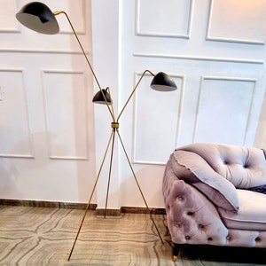 May include: A gold tripod floor lamp with three black lampshades. The lamp is in a room with white walls and a pink velvet sofa.