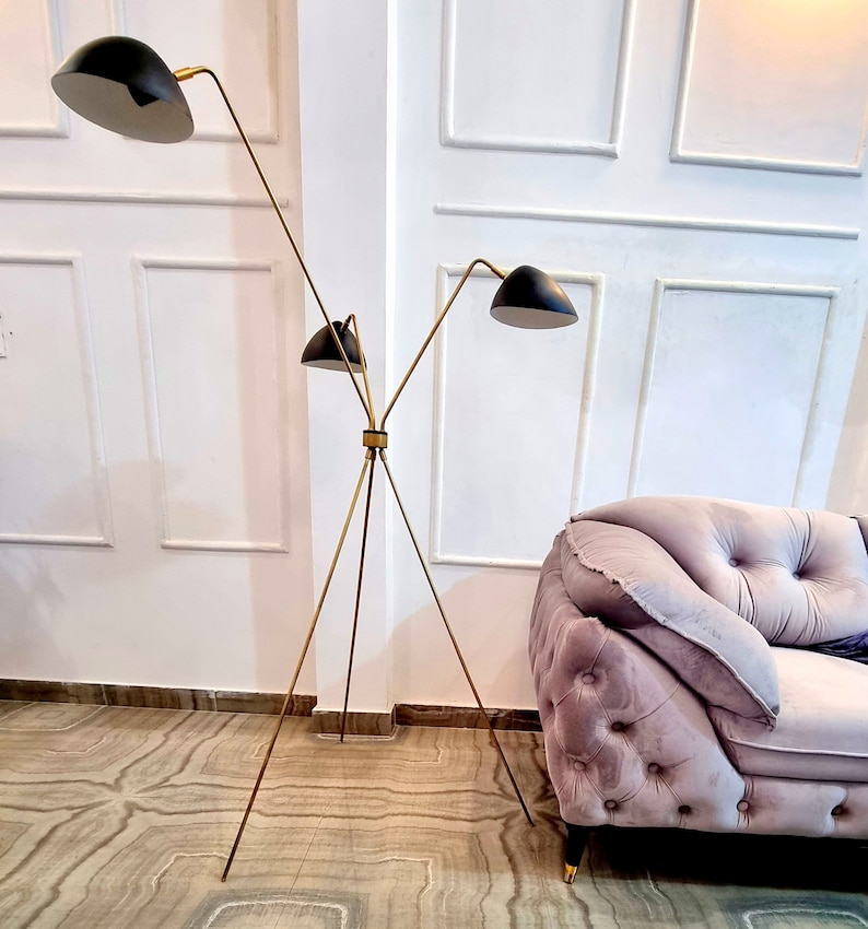 May include: A gold tripod floor lamp with three black lampshades. The lamp is in a room with white walls and a pink velvet sofa.
