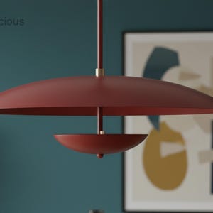 May include: A modern, red pendant light fixture with a large, flat shade and a smaller, bowl-shaped element below. The fixture hangs from a slender rod with a brass accent. The word "Chandelicious" is in the upper left corner.