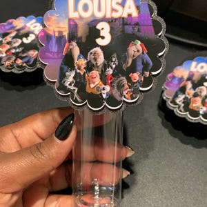 May include: A black and white circular favor with a scalloped edge. The favor features a colorful image of characters from the movie Sing 2. The text "LOUISA 3" is printed on the favor.