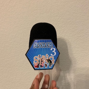 May include: A black microphone-shaped party favor with a blue and white hexagon-shaped label that reads "Asher 3" and "Sing" with cartoon characters. The favor is held by a person.