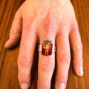 May include: A silver ring with a rectangular red enamel face featuring a gold figure and a black crown. The ring is on a finger, set against a wooden background.