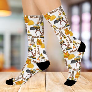 May include: White socks with a construction theme, featuring yellow hard hats, orange vests, and construction vehicles. The word "Matt" is printed repeatedly on the socks. The socks have black heels and toes.