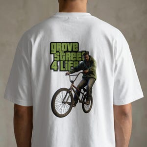 May include: White t-shirt featuring a graphic of a person riding a bicycle, with the text "GROVE STREET 4 LIFE" in green. The graphic depicts a person in a green hoodie and jeans. The shirt is made of a soft material.
