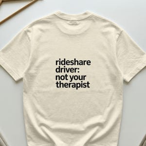 Op de afbeelding: Een crèmekleurig T-shirt met korte mouwen en zwarte tekst in het midden van de borst. De tekst luidt: rideshare driver: not your therapist. Het kledingstuk heeft een ronde hals en een standaard pasvorm met een zichtbare textuur.