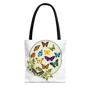May include: A white tote bag with black handles features a circular design of colorful butterflies on a cream background. The butterflies are arranged in a circle with green leaves and flowers around the edge.