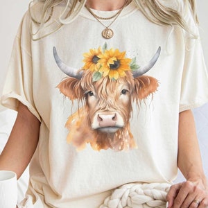 May include: A cream colored t-shirt with a watercolor illustration of a brown and white cow wearing a sunflower crown.