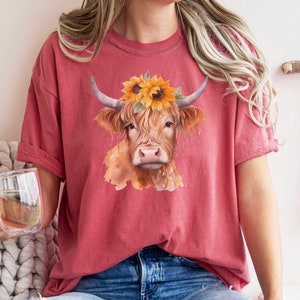 May include: A red t-shirt with a watercolor illustration of a Highland cow wearing a sunflower crown.