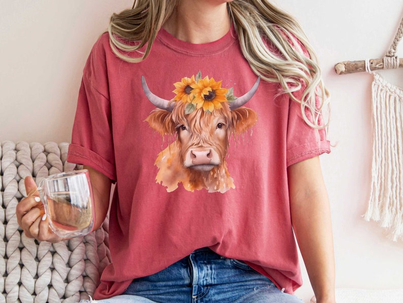 May include: A red t-shirt with a watercolor illustration of a Highland cow wearing a sunflower crown.