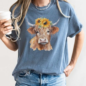 May include: A blue t-shirt with a watercolor illustration of a brown and white Highland cow wearing a sunflower crown.