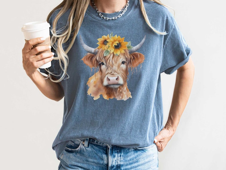 May include: A blue t-shirt with a watercolor illustration of a brown and white Highland cow wearing a sunflower crown.