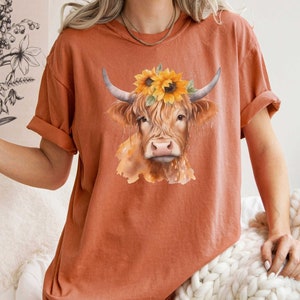 May include: A rust-colored t-shirt with a watercolor illustration of a brown Highland cow wearing a sunflower crown.