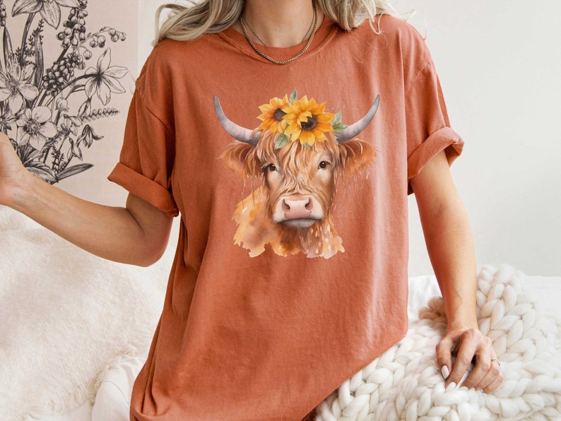 May include: A rust-colored t-shirt with a watercolor illustration of a brown Highland cow wearing a sunflower crown.