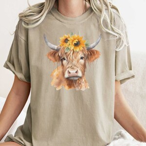 May include: A beige t-shirt with a watercolor illustration of a brown cow wearing a sunflower crown.