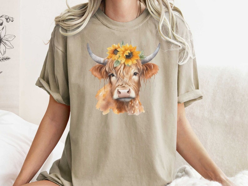 May include: A beige t-shirt with a watercolor illustration of a brown cow wearing a sunflower crown.